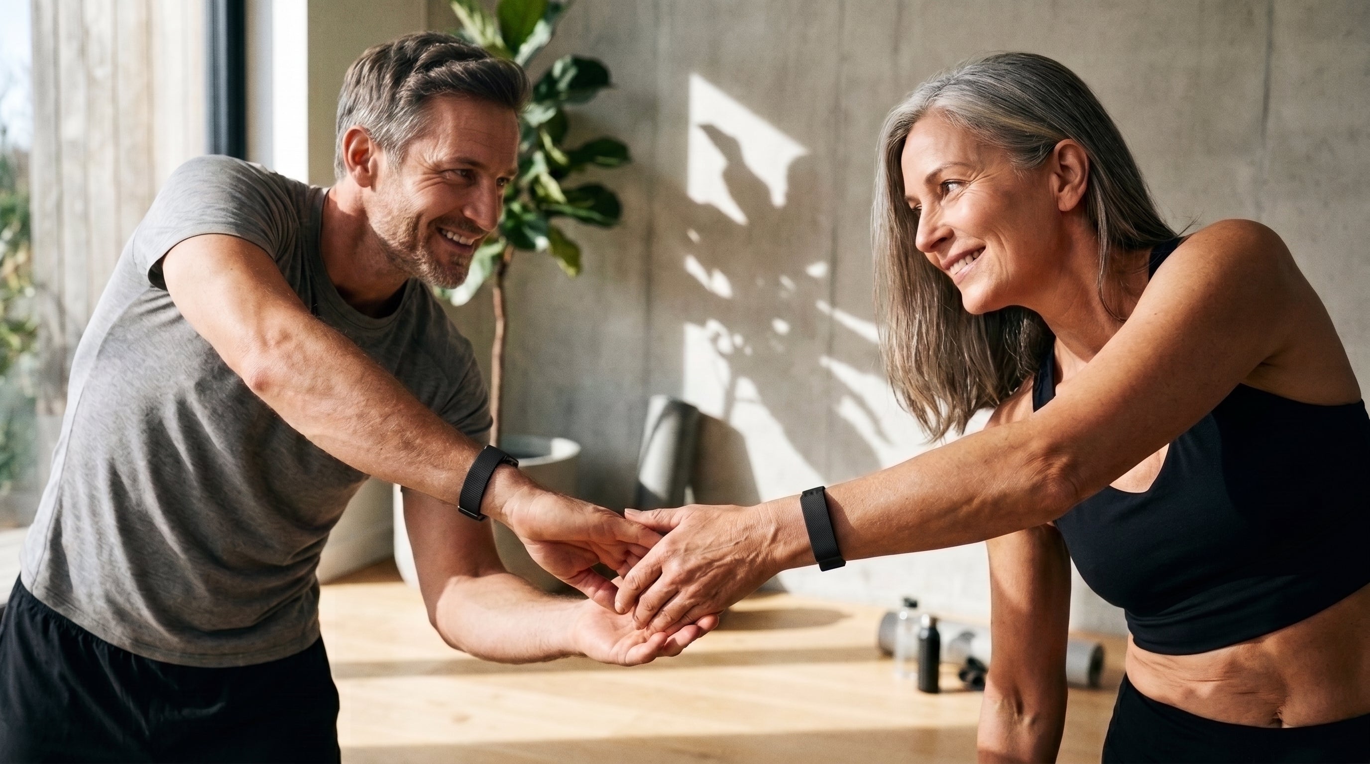 Two people smiling and clasping hands during a stretch at home, both wearing wrist bands